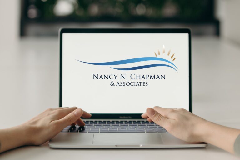 A counseling setting with a white woman typing on a laptop placed on a desk. The laptop displays the Nancy N. Chapman & Associates logo. A mirror is positioned behind the laptop, adding depth and a sense of reflection to the environment